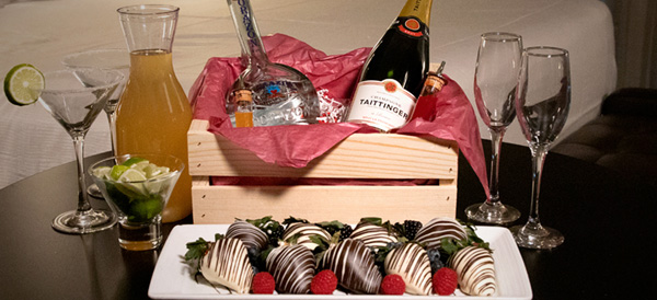 Table with chocolate covered strawberries plated in front of basket of tequila and champagne, mimosas, limes, champagne glasses, and martini glasses with salt rim.